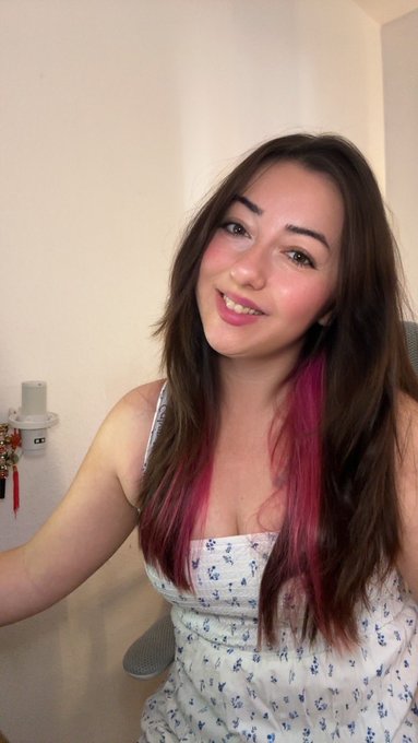 A young woman smiling in a selfie, wearing a white floral top, with long brown hair featuring vibrant pink highlights.