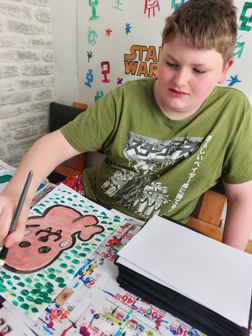 A boy in a green Star Wars t-shirt is painting a pink cartoon character in a sketchbook while seated at a table covered with art supplies.