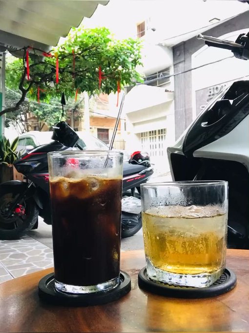 Two drinks sit on a wooden table at an outdoor café—a tall glass of iced coffee and a short glass of cold beer—while a motorbike and a car with an open trunk are visible on the street beyond. The scene captures a casual, reflective moment during a workday, fitting the post's message about taking things one day at a time while learning and building in the fast-moving Web3 space.