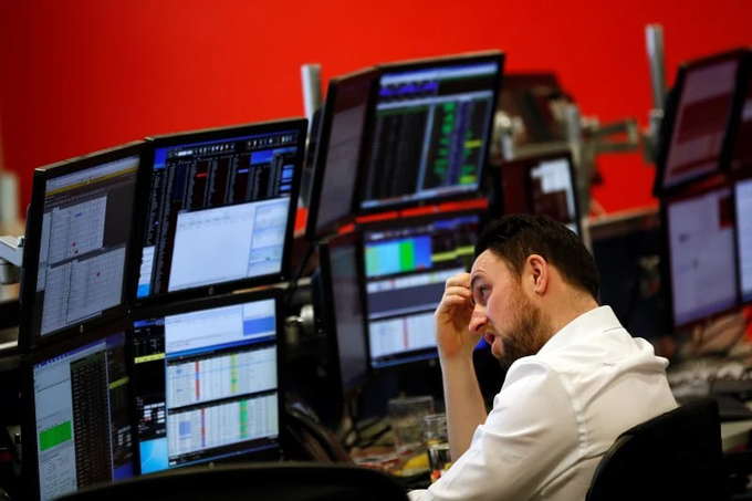 A trader in a white shirt sits at a desk surrounded by multiple computer monitors displaying financial data and trading screens, resting his head on his hand with a tired or stressed expression.