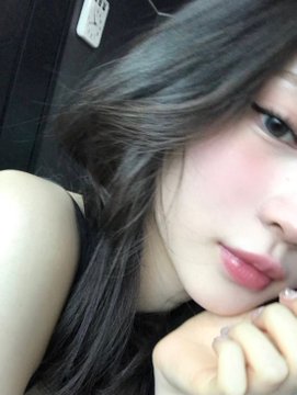 A close-up selfie of a young woman with long dark hair, prominent eyeliner, and glossy pink lips, resting her chin on her hand. No text or numbers are visible in the image.