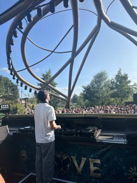 A DJ performs at the "LOVE" stage under a massive kinetic art installation at Tomorrowland in Belgium, with a large crowd visible beyond the booth. The word "LOVE" is prominently displayed on the DJ booth, tying into KuCoin's role as an official sponsor and exclusive Web3 partner of the festival.