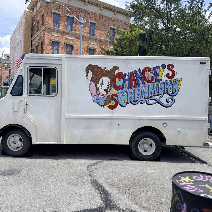 A white "Chance's Screamery" ice cream truck featuring a colorful clown logo is parked on a city street in front of a brick building with an American flag, suggesting the start of a relaxing vacation day with a sweet treat.