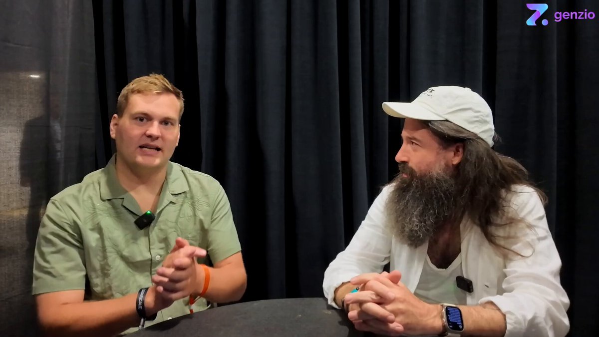 The image features two men sitting across from each other at a table, engaged in conversation. They are wearing hats and appear to be discussing something related to Web3 technology or blockchain. The background of the image shows a curtain, which could potentially be part of an event space or a meeting room where they are having this discussion.