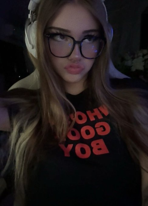 A young woman with long blonde hair wearing headphones, black glasses, and a black shirt with red "WHO GOOD BOY" text poses for a selfie, possibly showing the aftermath of someone "robbing" her of funds given her deadpan expression.