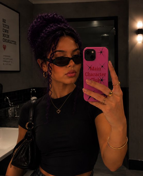 A woman with dark curly hair in a high ponytail, wearing black sunglasses and a black top, takes a mirror selfie with a hot pink phone case that reads "Main Character Club." The caption's "good morning web 3" ties into the bold, confident "main character" aesthetic of the image.