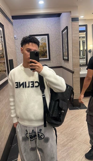 The image features a young man taking a selfie with his cell phone, wearing a white sweater that says "CELEAN" across the front. He has a backpack on his back and is standing in a bathroom stall or public restroom. The caption of the photo reads "New to X and am loving it #solana family over here is so massive I wish I was here years back".