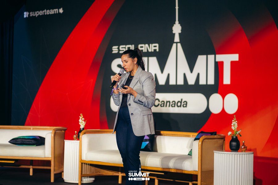 A woman in a gray blazer speaks into a microphone on stage at the Solana Summit Canada, with a red and black backdrop and a "superteam" logo.