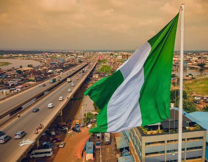A large Nigerian flag waves prominently in the foreground, with a highway bridge carrying traffic over a city landscape in the background.