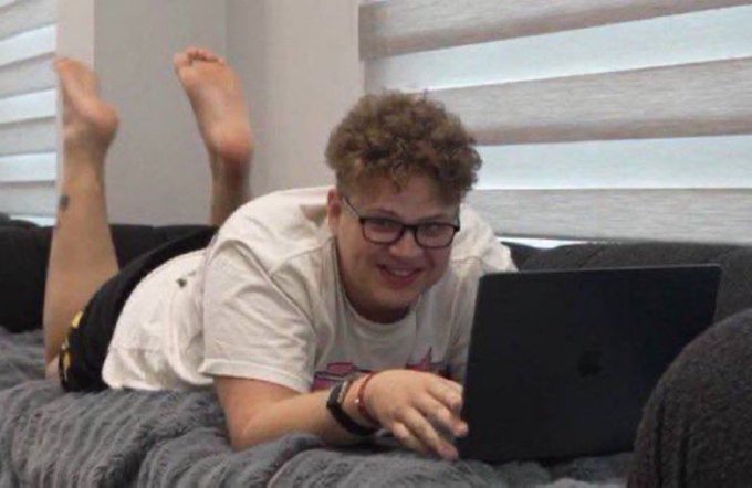 A person with curly hair and glasses lying on a bed, smiling while using a dark gray laptop.