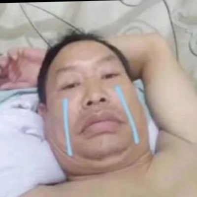 A man lies on a bed with his arm behind his head, with cartoon-style blue tears drawn on his cheeks to mimic crying.
