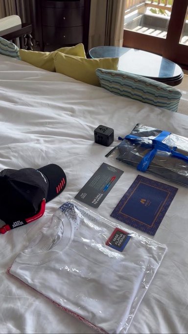 An assortment of merchandise items laid out on a hotel bed, including a black cap with red accents, a small black box, a dark booklet, an ornate blue and gold card or book, and a white t-shirt in plastic packaging.