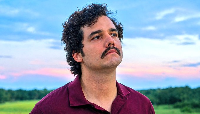 A close-up portrait of a man with curly dark hair and a mustache wearing a maroon polo shirt, looking upward against a backdrop of a pastel sunset sky.