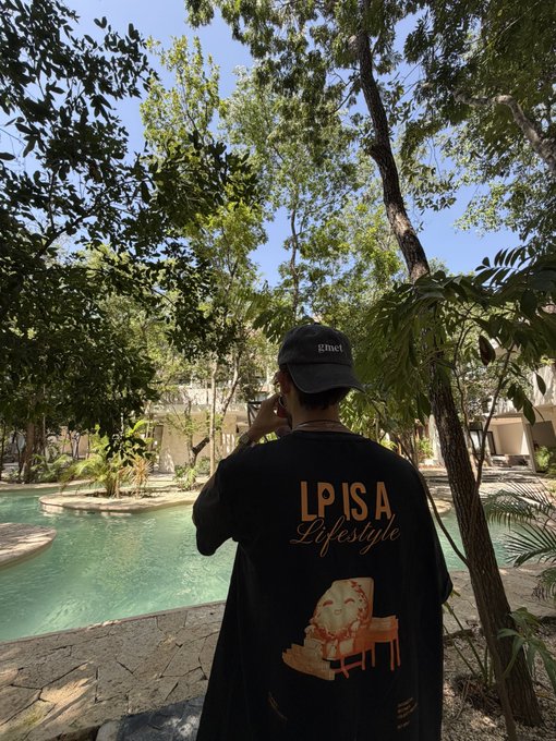 A person wearing a cap labeled "gmet" and a black shirt reading "LPISA Lifestyle" stands by a serene, tree-filled pool area, capturing the moment on their phone in response to a challenge from @MeteoraEco.