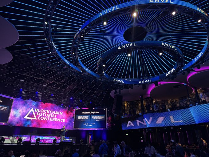 A large indoor conference venue featuring a striking circular LED ceiling installation displaying "ANVIL" repeatedly, with the "Blockchain Futurist Conference" logo projected on screens alongside a presentation titled "Buy Now, Pay Later," directly tied to the post about Anvil's discussion on how DeFi can perfect the BNPL concept. A speaker is visible on stage addressing the audience in this elaborate event space.
