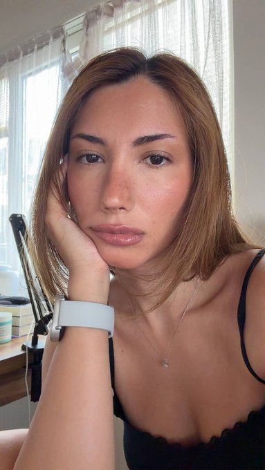 A close-up selfie of a woman with long light brown hair wearing a black top, a necklace, and a white watch, resting her chin on her hand indoors.
