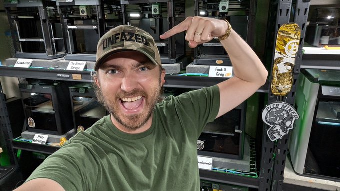 A man wearing a camouflage 'UNFAZED' cap and green t-shirt points at his head while posing in what appears to be a 3D printing workspace filled with various 3D printers.