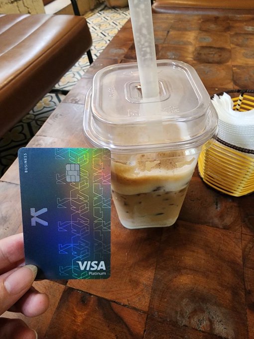 The image shows a person holding up a Visa credit card, which has a coffee drink inside of a clear plastic cup. The person's hand is positioned over the credit card, with the cup placed on top of the card. This scene suggests that the person might be using their Visa card to pay for the beverage or other purchases at a café or similar establishment.