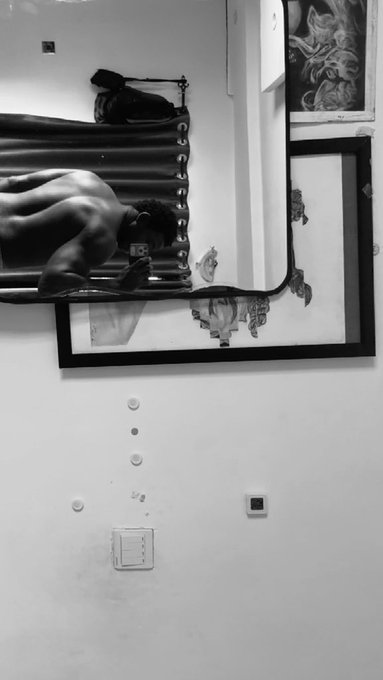 A black-and-white mirror selfie shows a shirtless individual with dark curly hair taking a photo in front of a framed artwork, with another piece of art visible in the upper corner. The image relates to the post's theme by depicting a casual "gm" (good morning) moment, capturing the subject seemingly preparing for the day ahead.