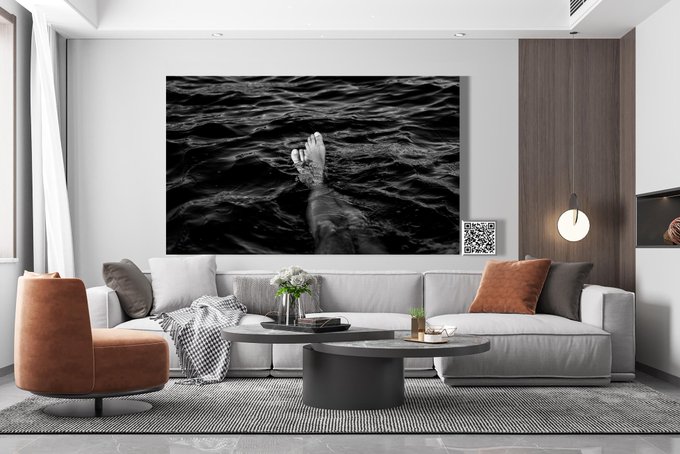 A modern living room featuring a large black-and-white photograph of a hand reaching out of dark water hanging above a gray sectional sofa with orange accent pillows.