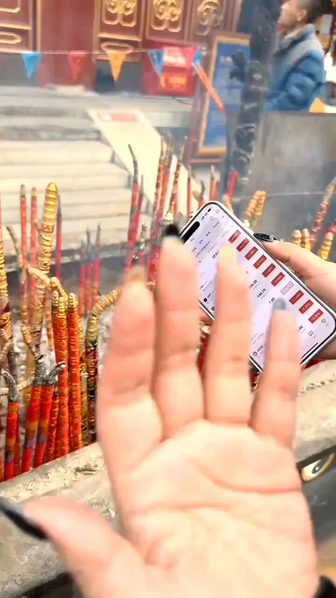 The image shows a person holding up their phone with a screen displaying a shopping list or a shopping app. The person's hand appears to be covering part of a temple wall, which has red and orange sticks arranged in rows. In the background, there are other people present who seem to be engaged in various activities.