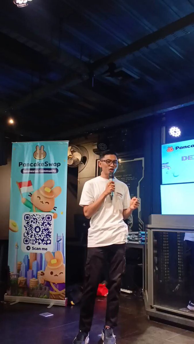 The image features a man standing in front of a banner that says "sharing session about pancake swap by PancakeSwap @BinanceAcademy #bandungdefi&web3". The man appears to be speaking into a microphone, possibly addressing an audience or participating in the event. There is also a poster visible in the background, which could provide more information about the pancake swap session or related activities.