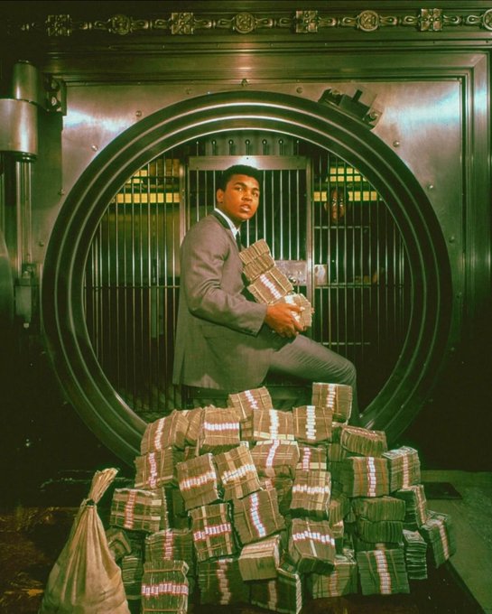 A man in a gray suit sits on a large pile of cash bundles inside an open bank vault.