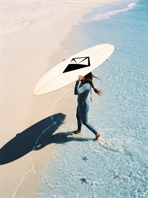 An aerial view of a surfer in a blue wetsuit walking along a sandy beach at the water's edge, carrying a white surfboard with a black geometric logo.