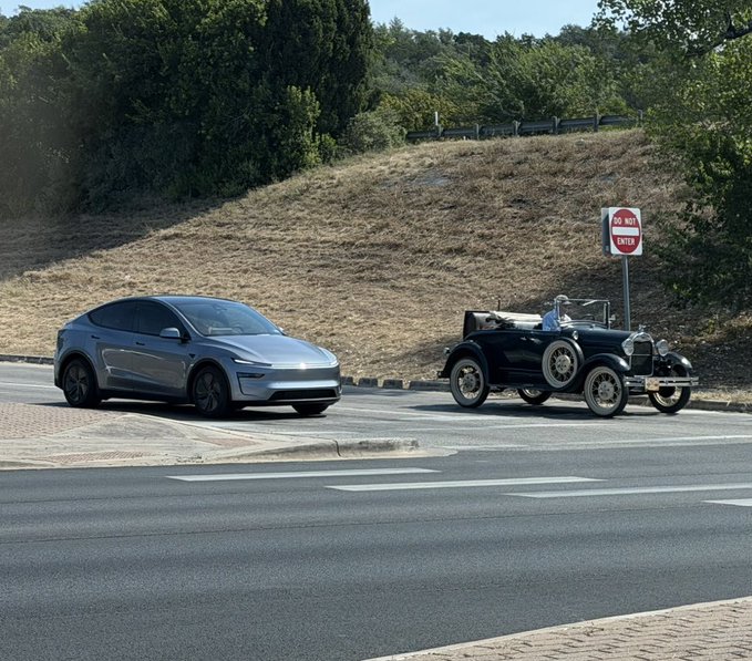 A modern silver Tesla is parked next to a vintage black classic car on a road near a 'Do Not Enter' sign, with a hillside in the background.