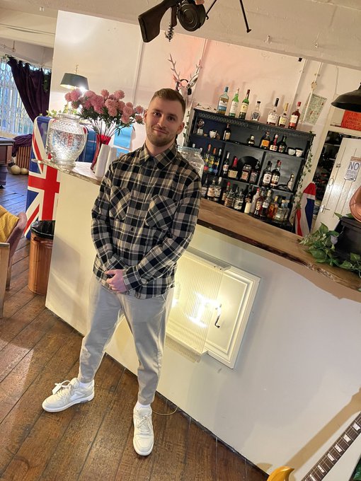 A man in a plaid shirt and gray jeans stands in a pub or bar setting, with shelves of liquor bottles, flowers, and Union Jack flags visible in the background.