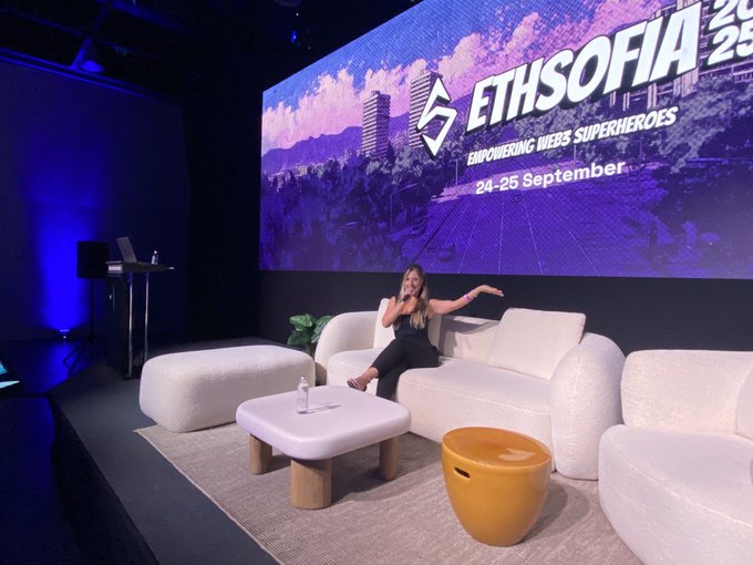 A woman sitting on a white couch on a stage presenting at ETHSofia, with a large screen behind her displaying 'ETHSOFIA - EMPOWERING WEB3 SUPERHEROES - 24-25 September'.