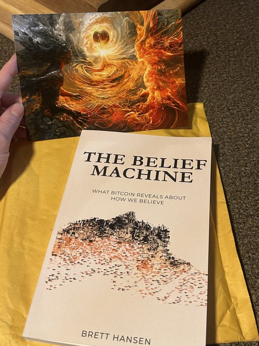 A person holds up a book titled 'THE BELIEF MACHINE: WHAT BITCOIN REVEALS ABOUT HOW WE BELIEVE' by Brett Hansen, alongside an artwork depicting a fiery, swirling abstract image.