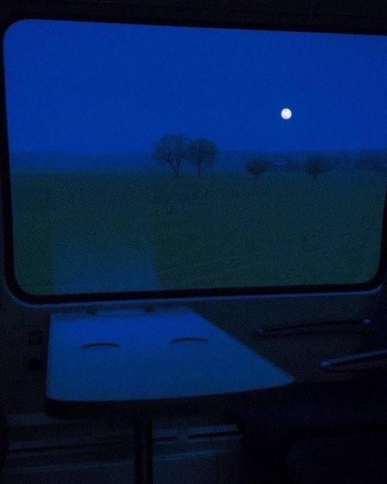 A moody photograph taken from inside a dimly lit train cabin, looking out the large window at a dark landscape with silhouetted trees under a deep blue night sky illuminated by a bright full moon.