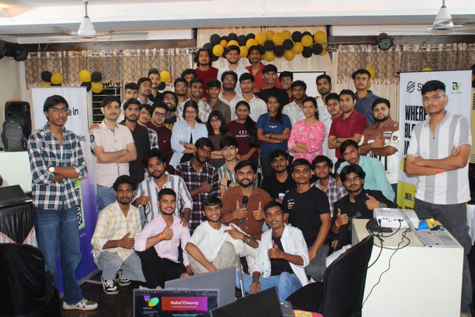 A large group photo of around 40 people poses at what appears to be a tech or crypto event, with banners for 'Sign' and 'Blockchain' visible alongside yellow and black balloons decorating the venue.