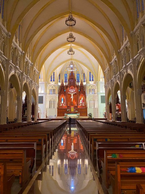 Interior view of an ornate cathedral featuring vaulted ceilings, hanging chandeliers, an elaborate wooden altar with religious statues, rows of wooden pews, and a highly polished reflective floor.