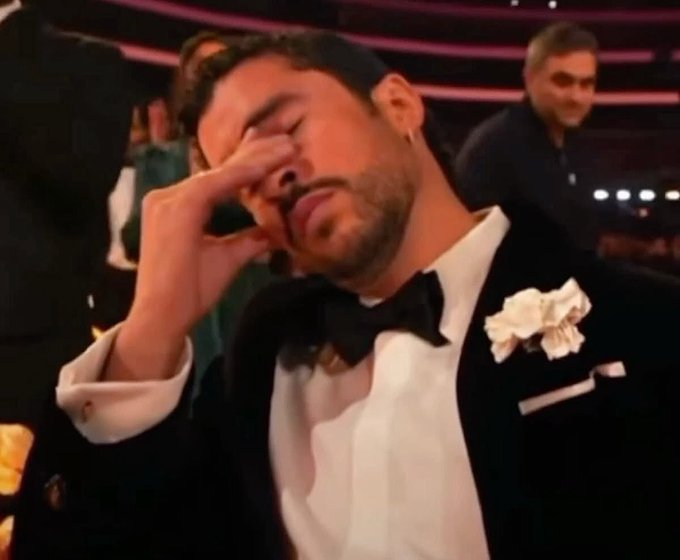 Crying man in a black tuxedo with a bow tie and white boutonniere, wiping tears from his eye, appearing to be at a formal awards ceremony event.