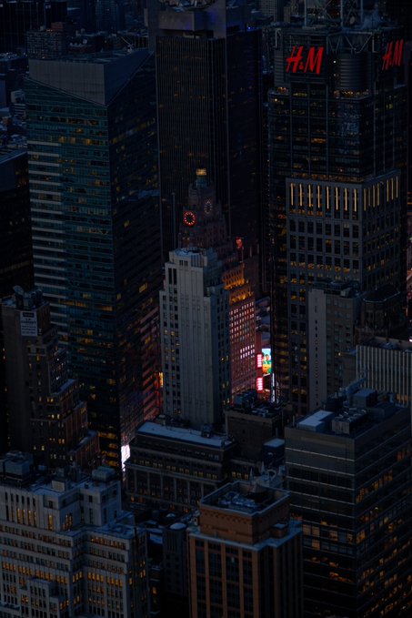 An aerial nighttime view of a dense cityscape, likely Times Square, with skyscrapers including the Paramount Building visible and an H&M sign in the background.