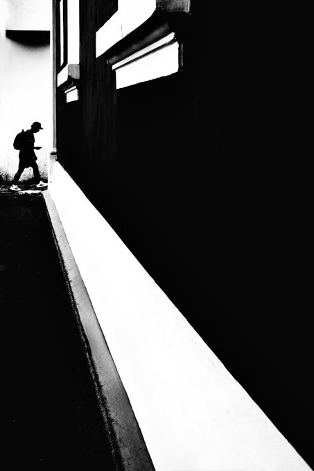 A high-contrast black and white street photograph showing the silhouette of a person with a backpack walking past a sharply angled building wall, creating strong diagonal lines and shadows.