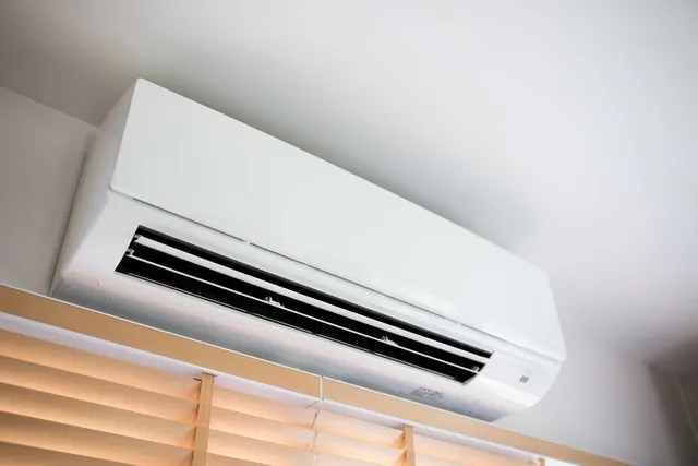 A white ductless mini-split air conditioner is mounted high on a wall above beige window blinds, viewed from a low angle. The image humorously underscores the "AC onchain" message, with the contraption—known for providing cool air—contrasted against the chaotic, "broken" energy of memecoin trading.