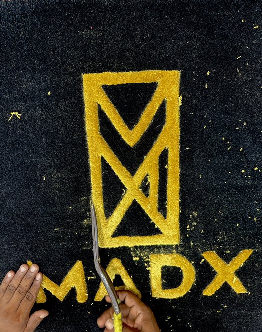A person's hand holds a small pickaxe near a yellow logo featuring an hourglass-like symbol with an "M" and "X" design above the word "MADX" painted or drawn on a black surface.