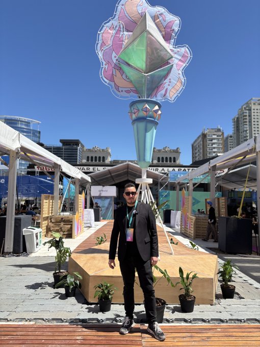 A man in a dark suit and sunglasses stands in front of a tall, diamond-topped monument or art installation at an outdoor event, with city buildings and tents visible in the background.