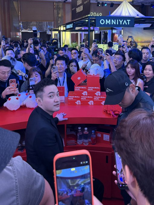 A slim man in black poses behind a red counter stacked with "TYRON BULL MUGS" boxes at a crowded event, surrounded by fans holding up phones to capture him, with "OMNIYAT" signage visible in the background. The image reflects the post's caption by showing the "Brother Sun" figure (likely crypto figure Sun Yuchen) at what appears to be a promotional event, capturing the energetic atmosphere of fans documenting his appearance.