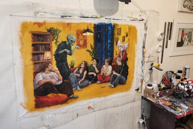 A large painting on a drop cloth in an artist's studio depicts an alien and various characters in a cozy yellow-walled room, surrounded by paint supplies and framed art.