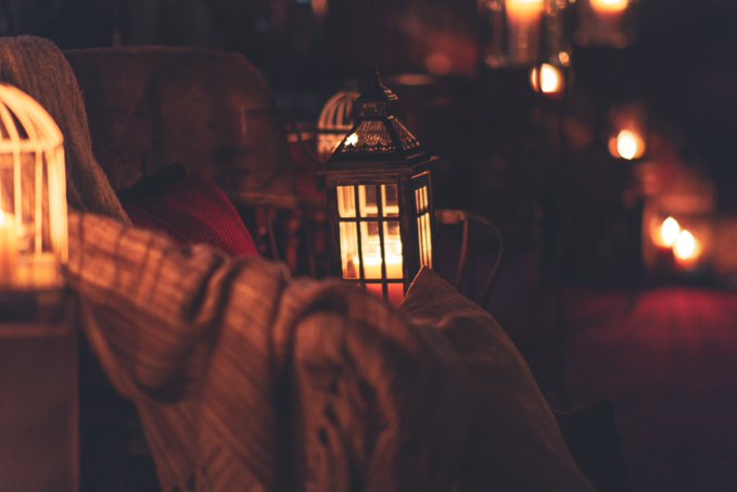 A dimly lit, atmospheric scene featuring multiple lit lanterns and candles, with a Buddha statue partially visible in the background behind draped fabric.