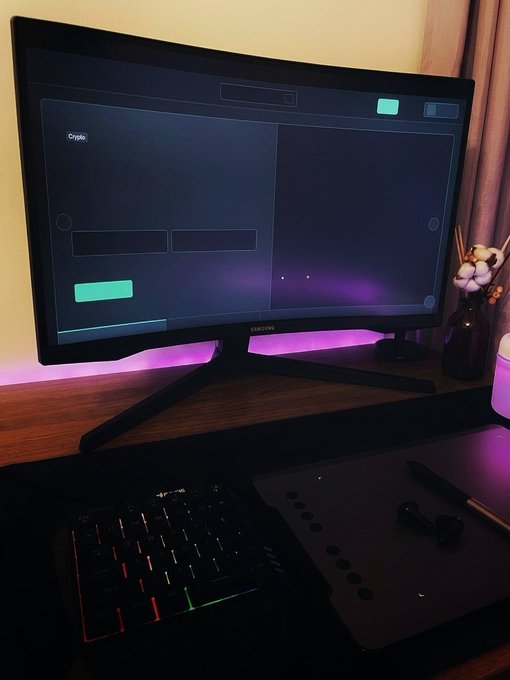 A curved monitor on a wooden desk displays what appears to be a game interface, with a backlit RGB keyboard and a graphics tablet on the desk, illuminated by ambient pink/purple lighting.