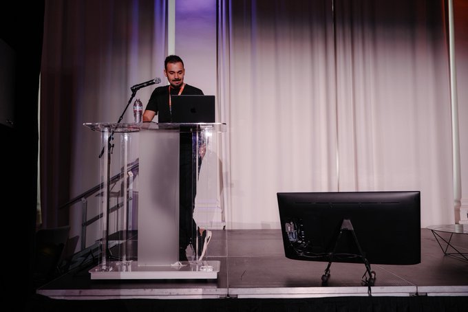 A man with a beard stands at a clear podium on a stage, speaking into a microphone with a laptop, while a monitor displays him.