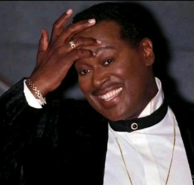 A smiling Black man in a black jacket with a white collared shirt touches his forehead with his hand adorned with a gold ring and bracelet.