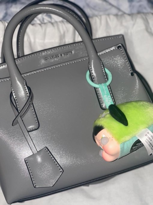 A grey leather handbag rests on a bed, adorned with a green plush toy keychain hanging from the handle.