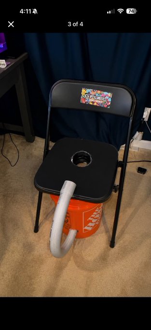 A black folding chair with a 'NO FUEL' sticker on the backrest sits over an orange Home Depot bucket, with a toilet paper roll placed in the seat's hole, creating a makeshift commode.