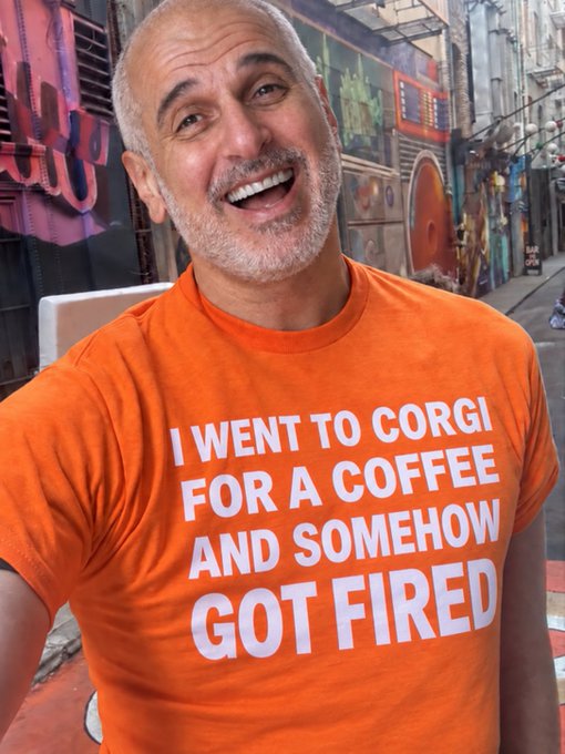 A smiling bald man with a gray beard wearing an orange t-shirt that reads 'I WENT TO CORGI FOR A COFFEE AND SOMEHOW GOT FIRED' stands in front of a colorful mural.